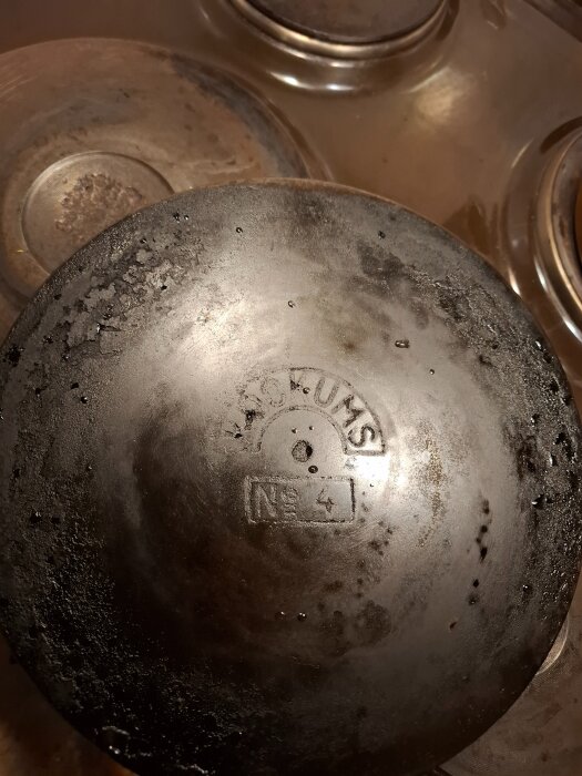 Sliten, gammal stekpanna med stämplad märkning, liggandes på spishäll. Från 95-årig morsas ärvda samling, möjligt att svetsa vid sprickor.