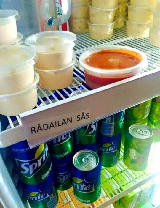Plastburkar med sås och läskburkar, bland annat Sprite, på en hylla i ett kylskåp märkt "Rådailan Sås".