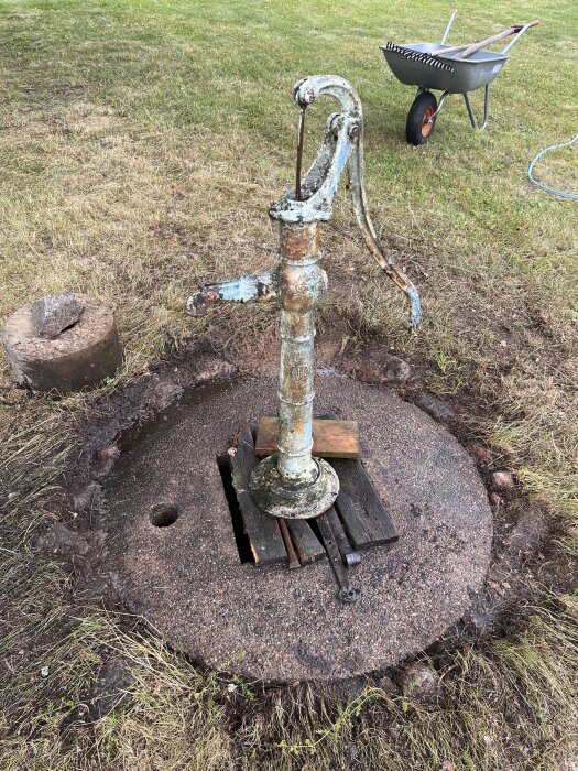 Gammal handpump monterad på betonglock över brunn, omgiven av gräs och jord, med en skottkärra i bakgrunden.