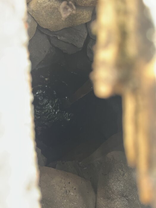 Gammal grävd brunn med stenar och vatten, sedd genom en öppning. Betongkant och träramar syns i förgrunden. Brunnen är delvis dold av skugga.