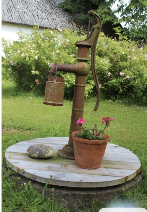 Gammaldags vattenpump på ett trälock ovanpå cementlock, med en blomkruka och sten som dekoration i trädgård.