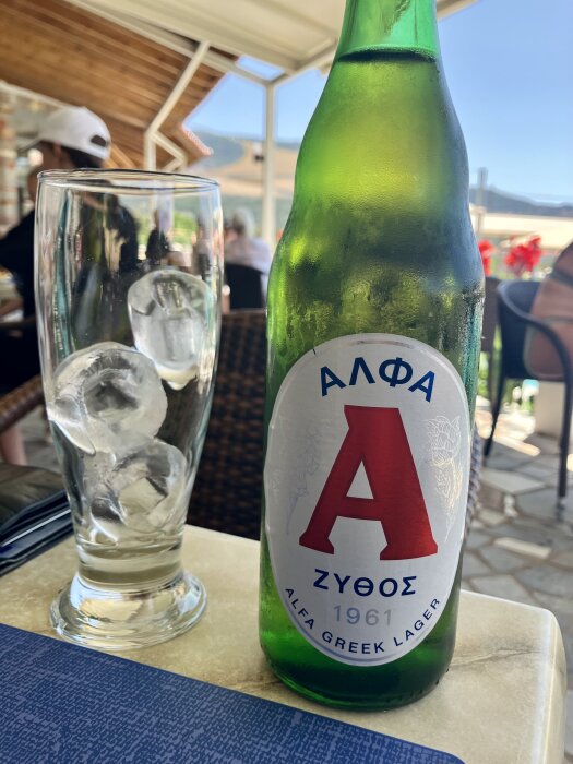 Grön Alfa Greek Lager-flaska bredvid ett glas med isbitar på ett bord i en restaurangmiljö.