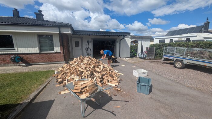 En tonårspojke staplar ved i en uppfart utanför en villa, bredvid en skottkärra full med ved och en släpvagn.