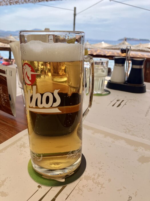 Ett glas öl med skum på toppen står på ett bord med ett havslandskap i bakgrunden.