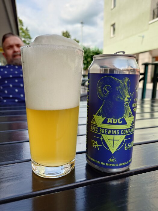 En burk Apex Brewing Company IPA vid sidan av ett glas med skummande öl på ett bord utomhus.
