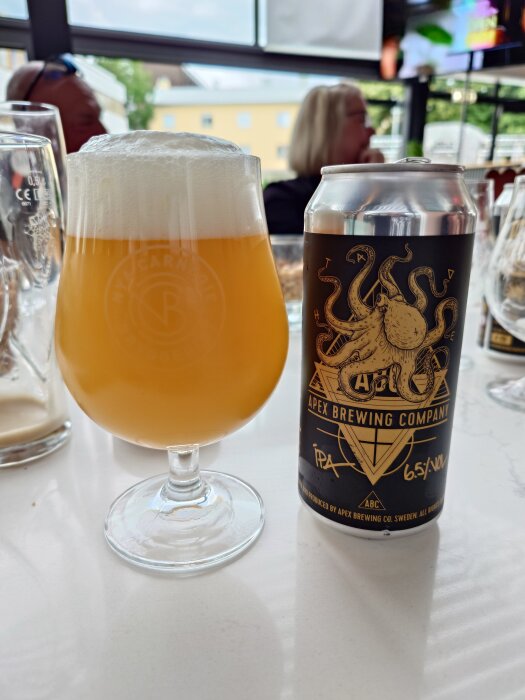 En glaskupa med skummande IPA och en burk från Apex Brewing Company visad på ett bord, med personer i bakgrunden.