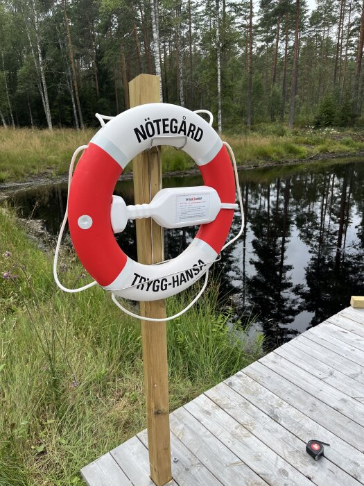 Räddningsboj märkt "Nötegård" och "Trygg-Hansa" vid en brygga med en skog och sjö i bakgrunden.