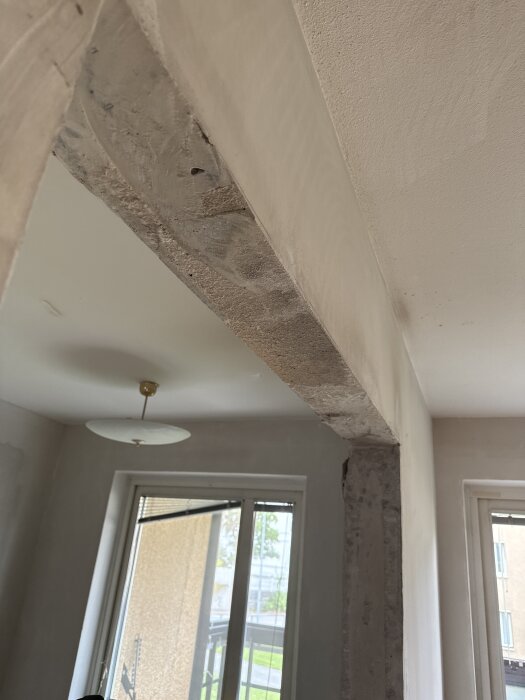 Öppning i en betongvägg med ojämna kanter, sedd inifrån ett rum med taklampa och fönster mot en balkong.