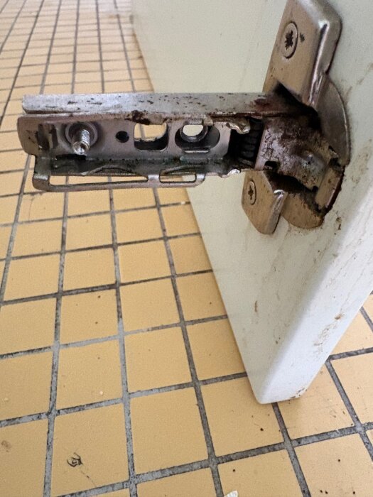 Närbild av ett köksskåpsgångjärn på en dörr, där en skruv har lossnat från fästet. Gult kakelgolv i bakgrunden.
