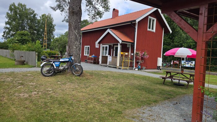 Rött caféhus med vita fönster, en parkbänk med parasoll och en motorcykel parkerad vid ett träd, omgiven av grönska.