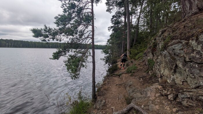 Person går på stig längs en sjö, omgiven av tallar och klippor. Molnigt väder.