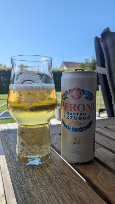 En ölglas och en burk Peroni Nastro Azzurro står på ett träbord utomhus, med blå himmel i bakgrunden.