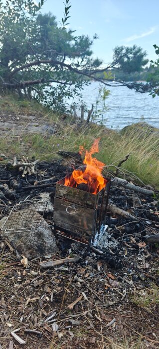 Portabel eldstad i metall med brinnande ved, placerad vid sjökanten i en skogsmiljö en klar dag.