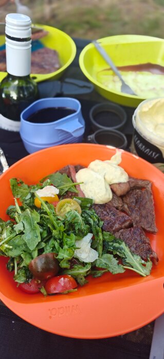 Grillad biff med bearnaisesås och sallad i orange tallrik, flaska och mugg i bakgrunden, utomhusmiljö.