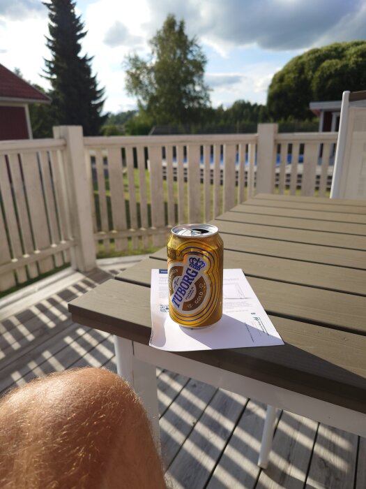En Tuborg ölburk på ett bord i solen på en altan med trästaket och gröna träd i bakgrunden.