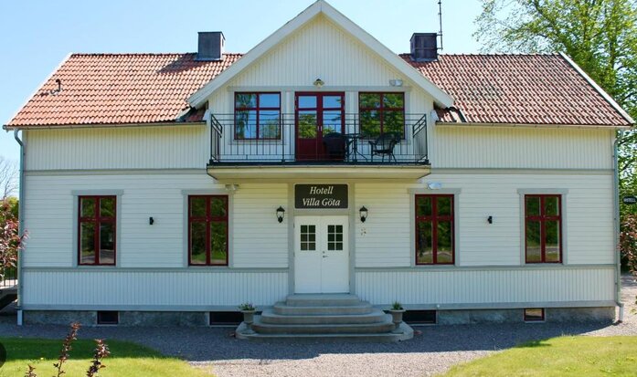 Vit tvåvåningsvilla med rött tegeltak och skylt "Hotell Villa Göta". Huset har röda fönsterkarmar och en liten balkong ovanför entrén. Grönska i bakgrunden.
