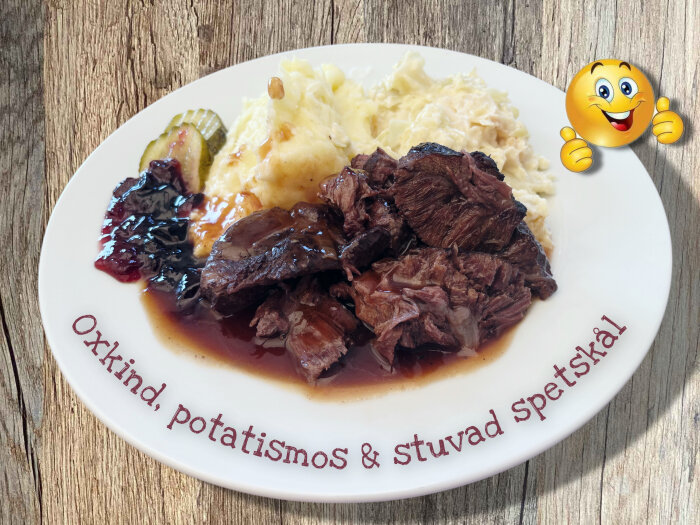 Tallrik med rödvinsbräserad oxkind, potatismos, stuvad spetskål, skysås, svartvinbärsgelé och inlagd gurka, med en glad emoji bredvid.