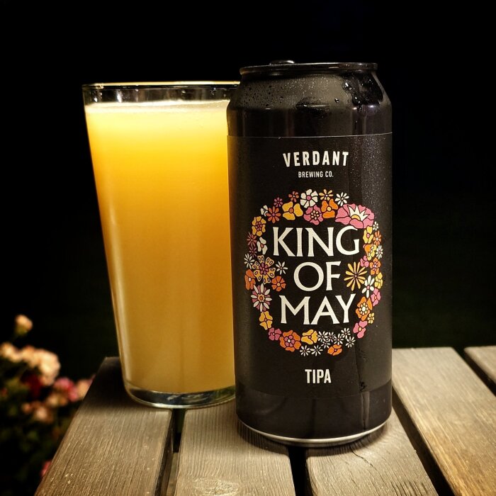 Bryggd burk med etiketten "King of May" bredvid ett glas ljus dryck på ett träbord.