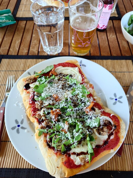 Pinsa med tonfisk, lök, tabasco, ruccola, balsamico, parmesan och picklade kantareller på tallrik, serverad med ett glas öl och vatten.