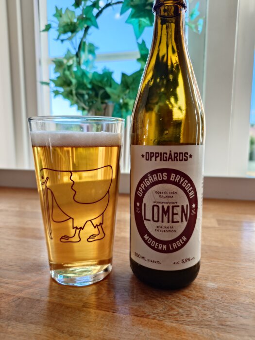 Flaska och glas med Oppigårds Lomen lageröl på ett bord, gröna växter i bakgrunden.