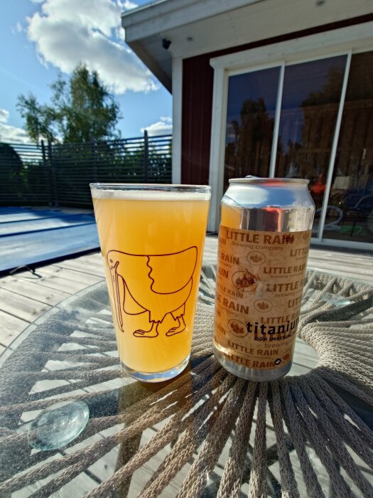 Ett glas med ljus öl bredvid en burk från Little Rain Brewing Company på ett bord utomhus under en blå himmel.