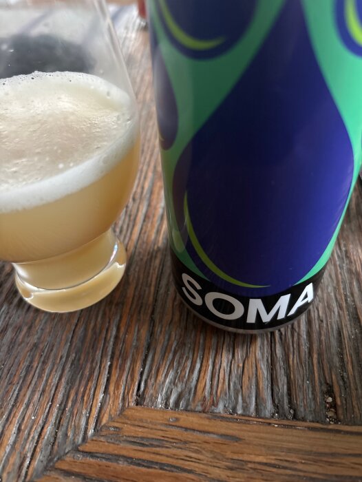 Ett glas med skummande dryck bredvid en färgglad burk märkt "SOMA" på ett träbord.