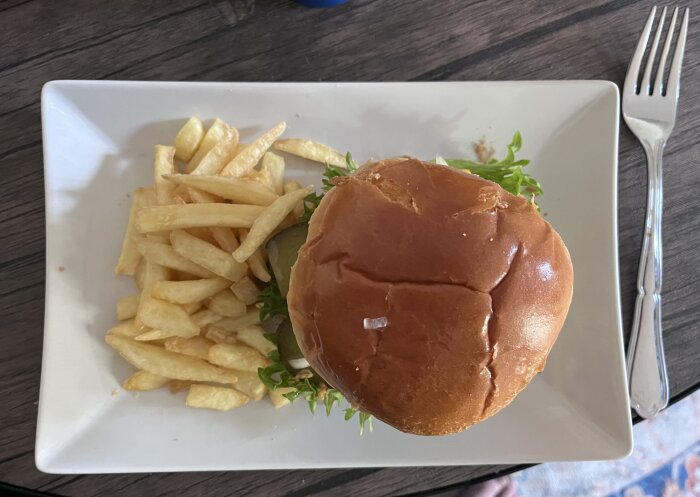 Hemlagad hamburgare med sallad, tomat och ost i bröd, serverad med pommes frites på en tallrik med gaffel bredvid.