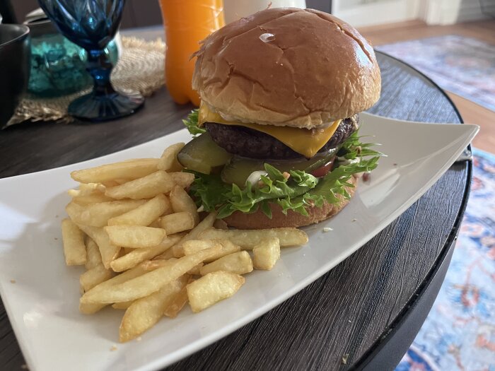 Hemlagad hamburgare med ost, lök, hamburgergurka, tomat, sallad och bröd, serverad med friterade pommes frites på vit tallrik.