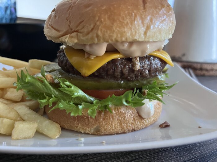 Hemlagad hamburgare med ost, sallad, gurka, tomat och dressing på en tallrik, serverad med pommes frites.