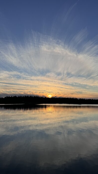 Solnedgång över en sjö med reflektioner i vattnet och träd i bakgrunden. Moln färgade av kvällsljuset mot en blå himmel.