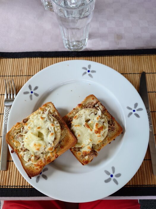 Två varma mackor med tonfisk, lök och ananas på en tallrik med blommönster, bestick och ett glas vatten i bakgrunden.