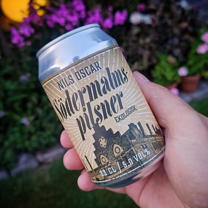 En hand håller en burk Södermalms Pilsner, ekologisk öl, med en bakgrund av suddiga blommor och trädgård.