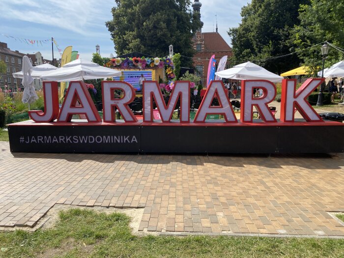 Stora röda bokstäver bildar ordet "JARMARK" på ett utomhusevent med blommor och parasoller i bakgrunden.