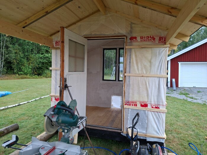 Litet trähus under byggnation, med öppen dörr och grundmålat interiör. En cirkelsåg står framför. Gräs och en röd byggnad syns i bakgrunden.