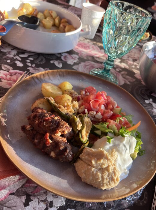 Grekisk middag utomhus med köttfärsspett, ugnstekt potatis, vattenmelon- och fetaostsallad, hummus, tzatsiki och fyllda padrones på tallrik.