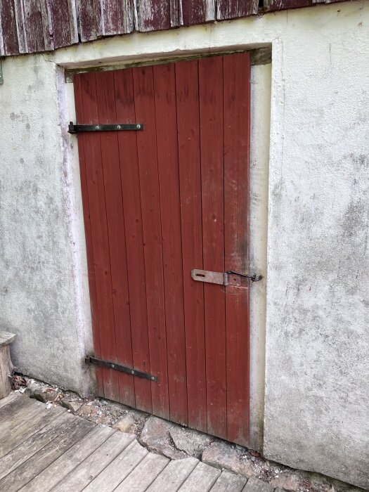 Röd enkel dörr med gångjärn och hasp, monterad i en vit vägg, vid en träveranda.