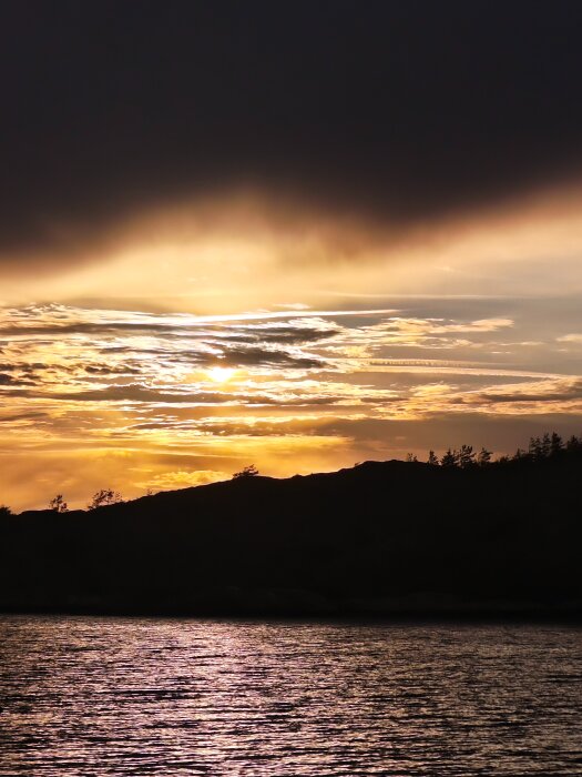 Solnedgång över havet med dramatisk himmel och siluett av trädtoppar i bakgrunden.