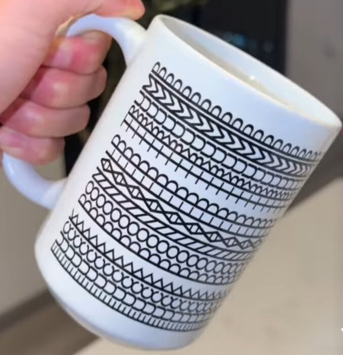 <!-- En hand håller en vit mugg med svart geometriskt mönster. -->