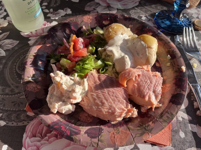 Tallrik med lax, kokt potatis med sås, sallad och skagenröra på blommigt bord.