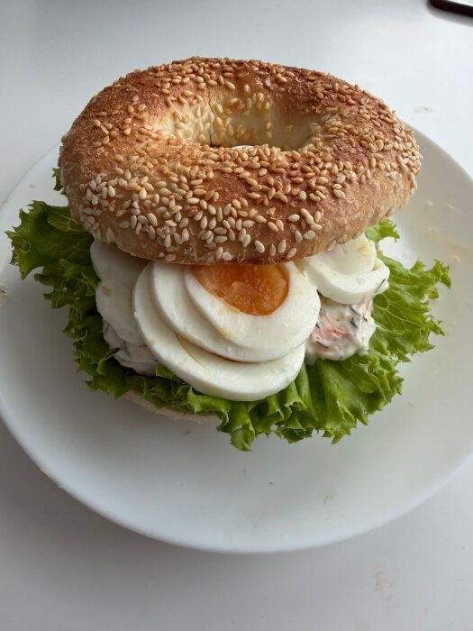 Sesambagel fylld med räkröra, skivade ägg och sallad på en vit tallrik.