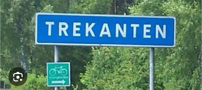 Vägskylt med texten "Trekanten" framför grönskande bakgrund. Under finns cykelskylt med pil åt vänster.