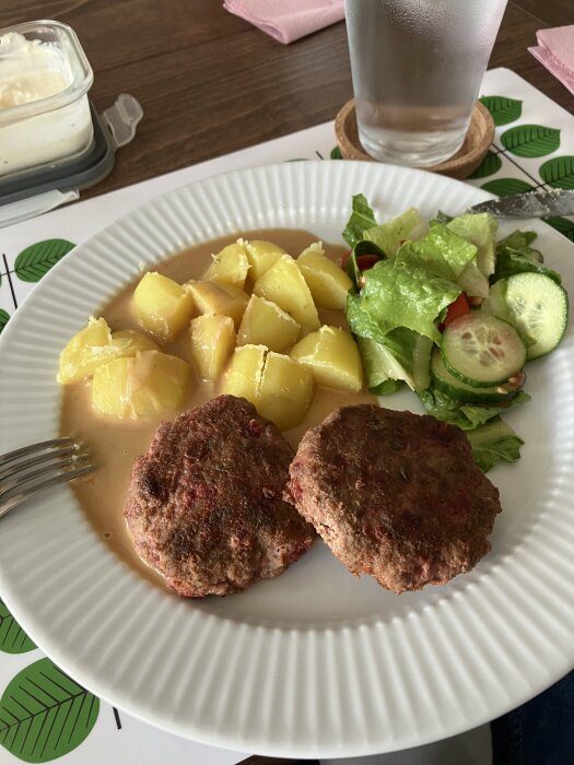 Tallrik med nötfärsbiffar med rödbetor, potatis i sås och grönsallad, serverad på vit tallrik med ett glas vatten bredvid.