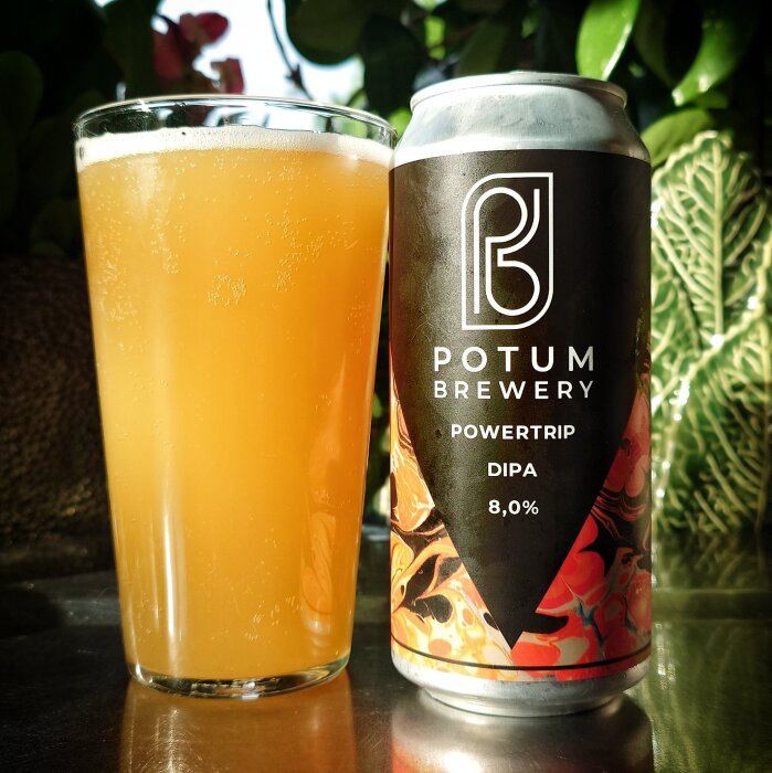 Ölglas med ljus, grumlig dryck bredvid en burk Potum Brewery Powertrip DIPA, 8% alkoholhalt, med växter i bakgrunden.
