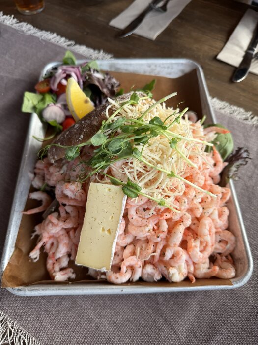 Räkmacka med bröd, ost, räkor, groddar och citron på en bricka, serverad på en dukad träbord.
