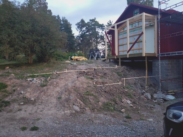 Pinnar markerar plats för nya stödmurar vid en lada under ombyggnad. I bakgrunden syns grönska och en del av byggnaden på pelare.