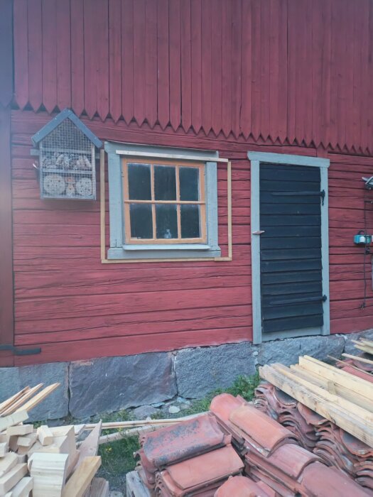 Röd ladugårdsvägg med ett fönster, dörr och staplade byggmaterial på marken.