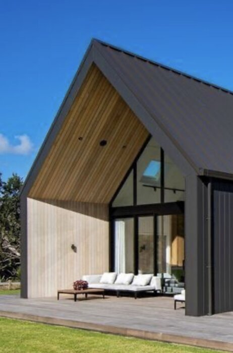 Modern villa med snedtak och stora glasdörrar, uteplats med soffor och bord, omgiven av grön gräsmatta och blå himmel i bakgrunden.