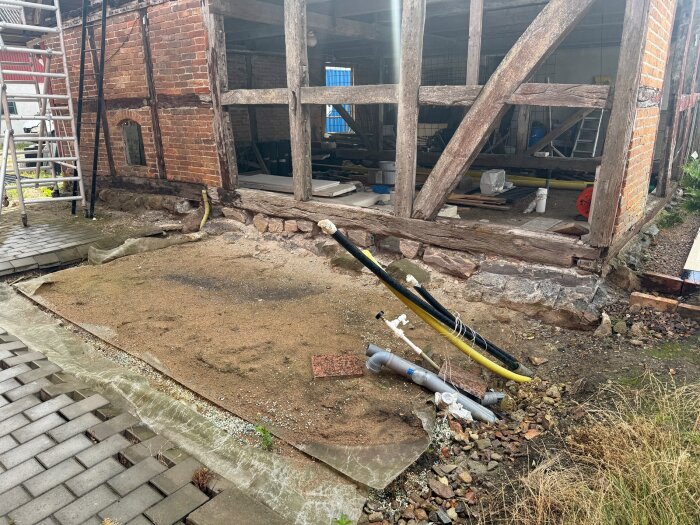 Exponerad fasad av tegel och trä hos en renoveringsbyggnad. Grävd mark synlig med avloppsrör, omgiven av en stenlagd yta och vegetation.