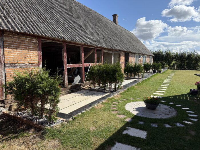 Tegelhus med sadeltak och trädgård, omgiven av grönska och stenlagd gång. Buskar kantar stigen och blomkrukor pryder gräsmattan.