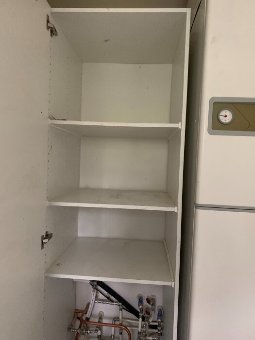 Öppet skåp med hyllor, nederst syns rör och en vattenmätarkonsol monterad. Värmeenhet syns till höger.